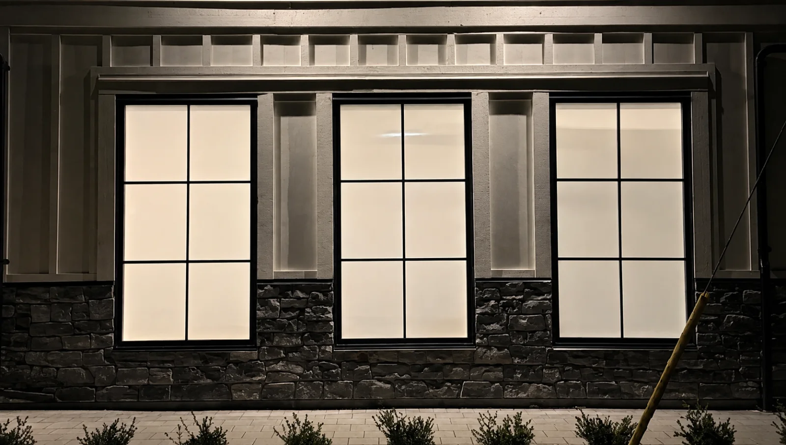 A home exterior at night with three windows glowing through roller shades
